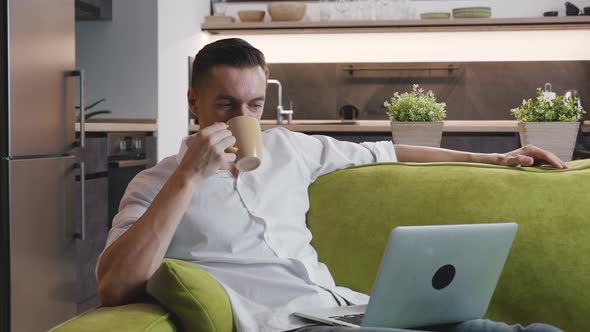 Smiling Young Man Freelancer Is Relaxing on a Cozy Couch, Drinking Delicious Coffee and Watching