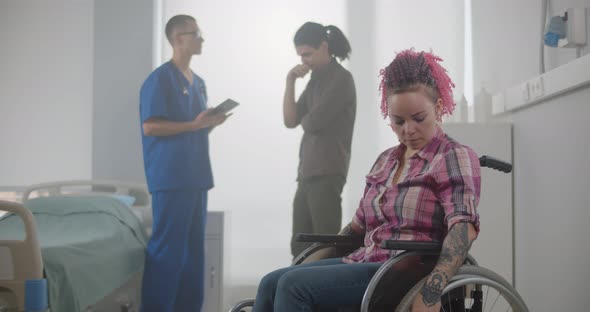 Upset Disabled Young Woman in Wheelchair with Doctor and Husband Talking on Background