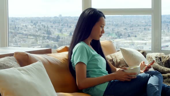 Woman using mobile phone while having breakfast in living room