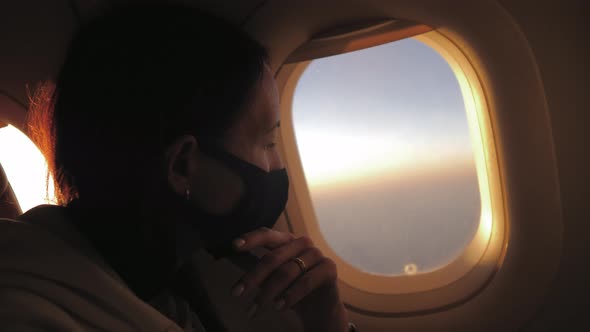 Young Woman Sits on a Plane in a Black Medical Respiratory Mask and Looks Out the Window