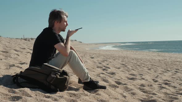 Young Man Sitting on Coastline Having Conversation with Voice Message Recorder on Smartphone