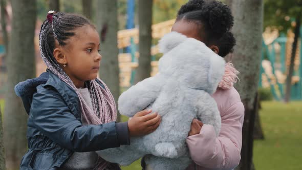 Girls Looking at Camera Two Friends Sitting on Bench in Park Kid Gives Teddy Bear Little Sisters