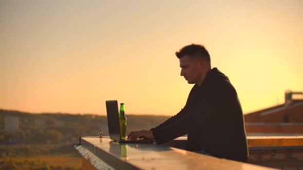 A Male Freelance Programmer Sits on a Skyscraper Roof with a Laptop and Beer Typing Code 