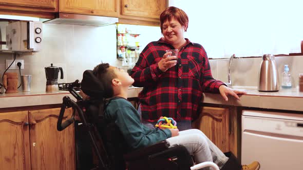 Latin mother taking care of teen son with disability on wheelchair inside home kitchen