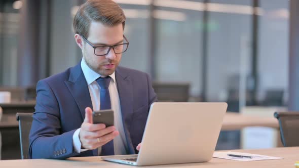 Businessman with Laptop Browsing Internet on Smartphone at Work