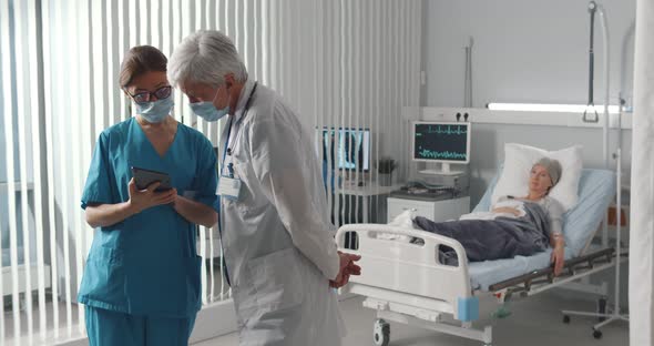 Doctor and Nurse in Safety Mask Discussing Diagnosis on Tablet with Woman Patient Lying in Bed