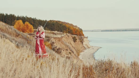 Blonde Woman Warrior in Red National Dress Standing on the Mountain Cliff and Wields a Sword