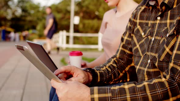 Man Uses an Electronic Tablet Against Background of Woman with Laptop and Coffee Closeup Against