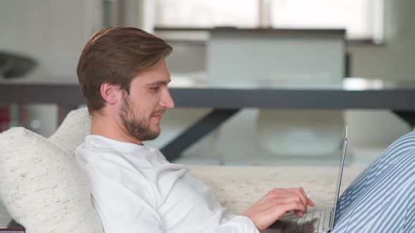 Cheerful Caucasian Man Sits at Home on the Couch and Works Using a Laptop Manager on a Remote Work