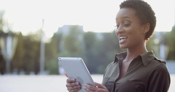 Young Ethnic Woman Wears Shirt, Stands on Street on Background of Sunset, Uses Computer Tablet for