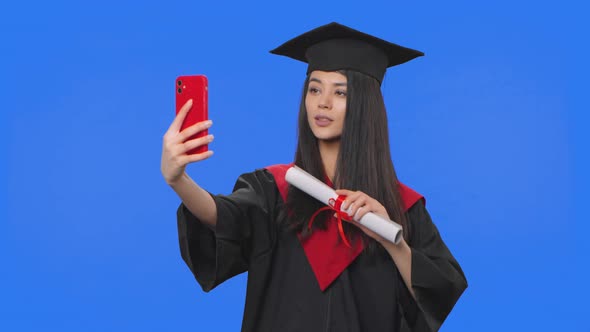 Portrait of Female Student in Graduation Costume Holding Diploma and Making Selfie on Mobile Phone