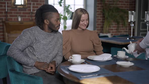 Positive Rich Couple Talking Sitting in Restaurant As Unrecognizable Waiter Pouring Tea From Teapot