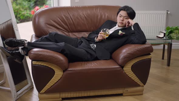 Desperate Frustrated Young Asian Groom Lying on Armchair Holding Glass of Whiskey