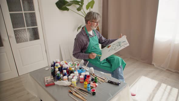 Man artist paints on canvas sitting on chair in an apron stained with paints, dipping brush into jar