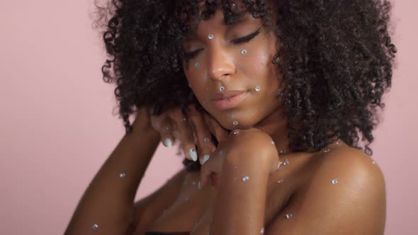 Mixed Race Black Woman with Curly Hair Covered By Crystal Makeup on Pink Background in Studio