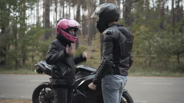 Angry Couple of Biker Arguing Outdoors. Irritated Young Woman Taking Off Pink Helmet, Giving It To