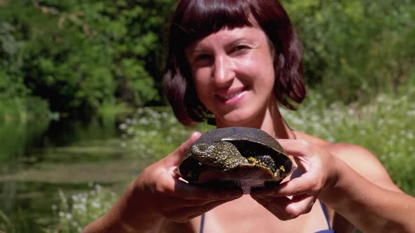 Woman Holds Funny Turtle in Arm and Smiles on River with Green Vegetation