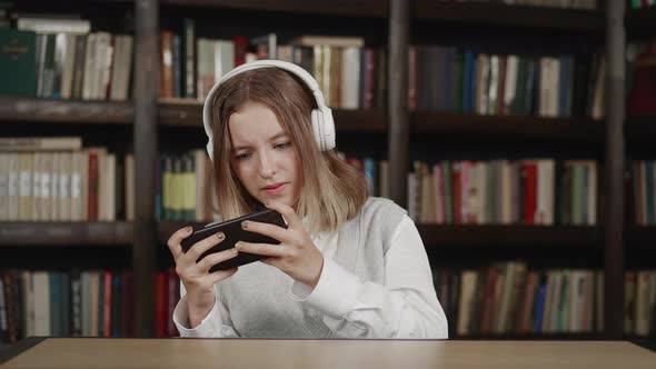 Lazy Schoolgirl with White Headphones Smartphone Distracting From Homework Sitting in Library