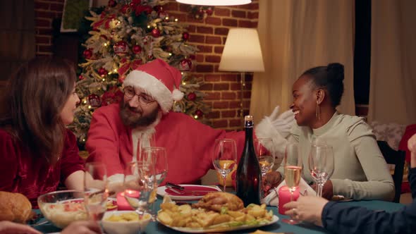 Festive Man Wearing Santa Claus Costume at Christmas Dinner Party Talking with Family Members