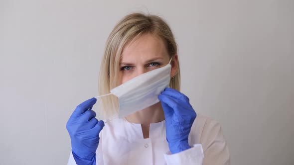 Blonde Female Doctor Surgeon in White Uniform and Blue Gloves Puts on Medical Mask