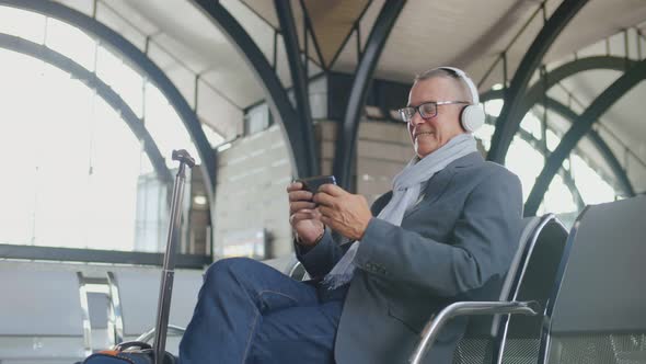 Aged Businessman Using Phone and Headphones Waiting for Delayed Flight at International Airport