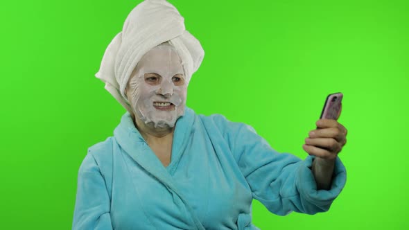 Grandmother in Bathrobe, Face Mask. Old Woman Making Selfies Using Mobile Phone