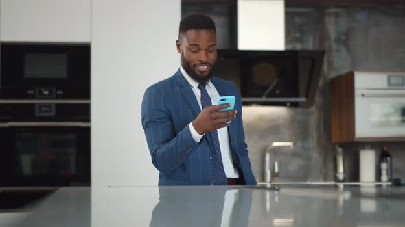 Smiling African Businessman Wearing Suit Receiving Good News on Cellphone at Modern Kitchen