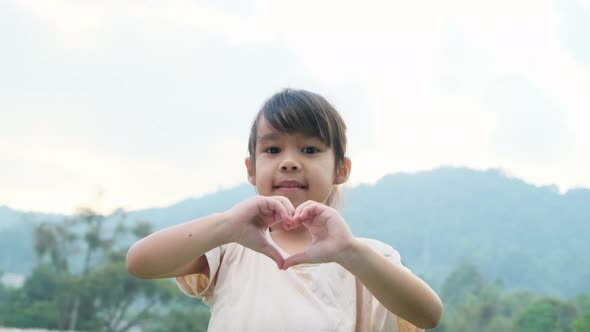 Cute Asian preschooler smiling happily and making heart-shaped hands on green nature background.