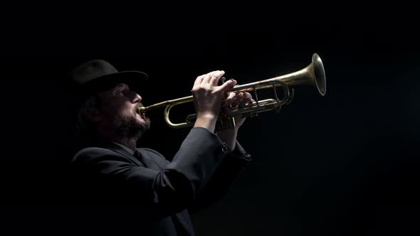 Man in jacket stands under spotlight in dark studio and plays classical piece on trumpet, side view.
