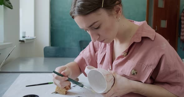 Female Potter Sitting and Stirs Paint with a Brush a Cup on the Table. Woman Making Ceramic Item