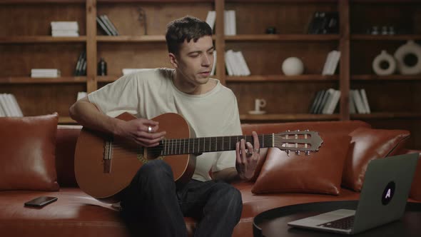 20s Man Learning To Play the Guitar Looking at Laptop Sitting on the Sofa at Home