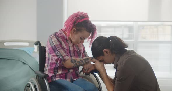 Portrait of Sad Young Man Kneeling Before Girlfriend in Wheelchair at Hospital