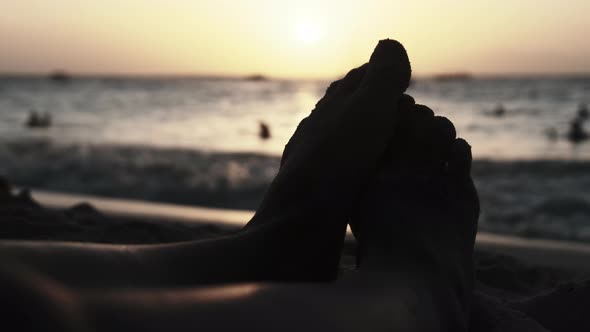 POV Silhouette of Feet of Young Man Lying on Sandy Beach By Ocean During Sunset