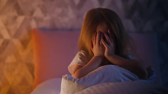 Unhappy Child Hides Closing Eyes with Palms on Bed in Room