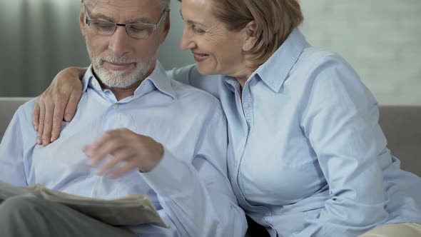 Elderly man on couch reading newspaper, woman sitting beside hugging him, caring