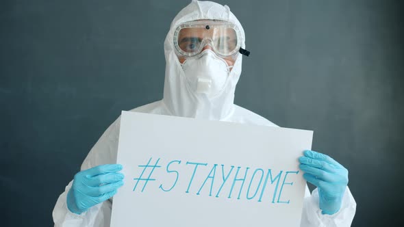 Portrait of Arab Man in Quarantine Suit Standing with Stayhome Banner on Grey Background alt