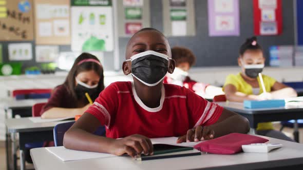 African american boy wearing face mask while sitting on his desk in the class at school