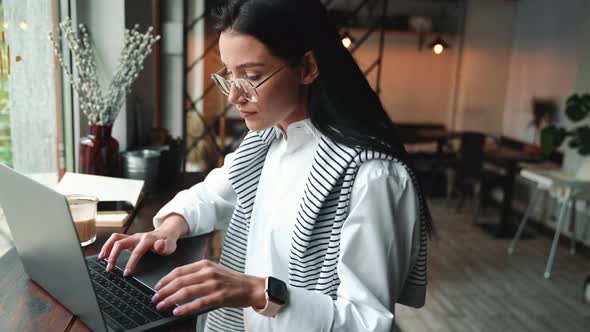 Serious young brunette woman in eyeglasses working on laptop