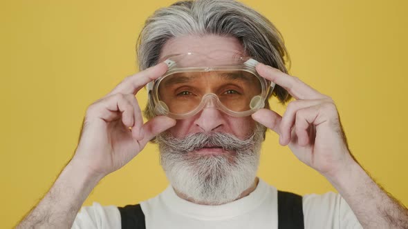 Senior Handyman Putting on Protective Glasses on Yellow Background