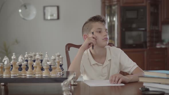 Portrait of a Pensive Little Boy Sitting at the Table at Home. The Guy Is Holding a Pen Thinking