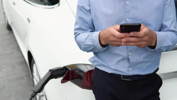 Close Up Man Hand Holding a Smartphone Near Charging Electric Car EV Car