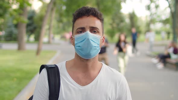 A Young Handsome Man Stands in the Park in the Alley Wearing a Protective Mask During the