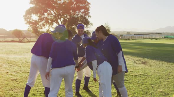 Diverse group of female baseball players on sunny pitch, in a huddle, stacking hands