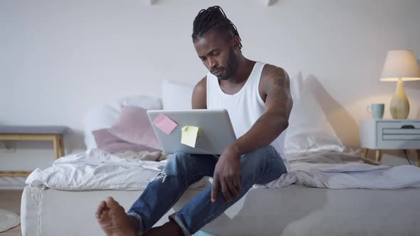 Wide Shot Portrait of Overwhelmed African American Tired Man Sitting on Bed with Laptop Holding Head