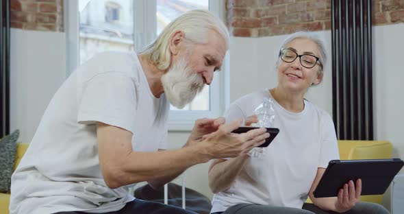 Mature Couple in white T-shirts Having Fun Together while Revisioning Funny Videos on Mobile 