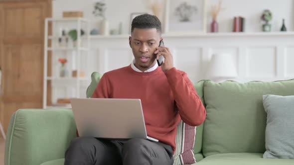 Angry African Man with Laptop Talking on Phone on Sofa