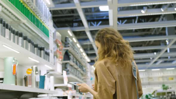 Beautiful Woman Chooses a Hot Mug in a Mall with a Tablet Computer Pc, a Shopping List in Electronic