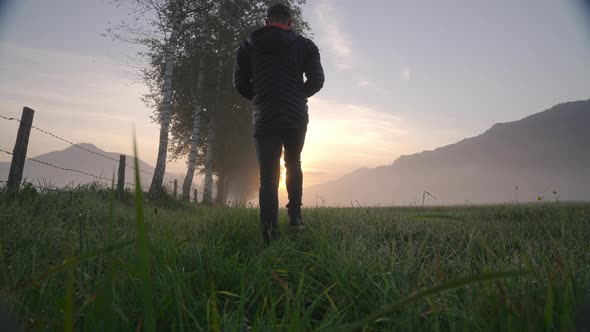 Man Walking Through Meadow To Foggy Landscape And Sunrise