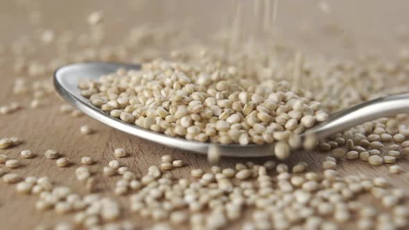 Raw quinoa seeds fall into a metal spoon in slow motion on a wooden board