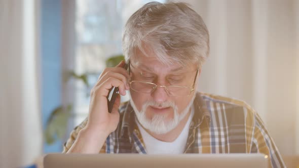 Concerned Senior Man Talking on Mobile Phone Sitting at Desk Looking at Laptop